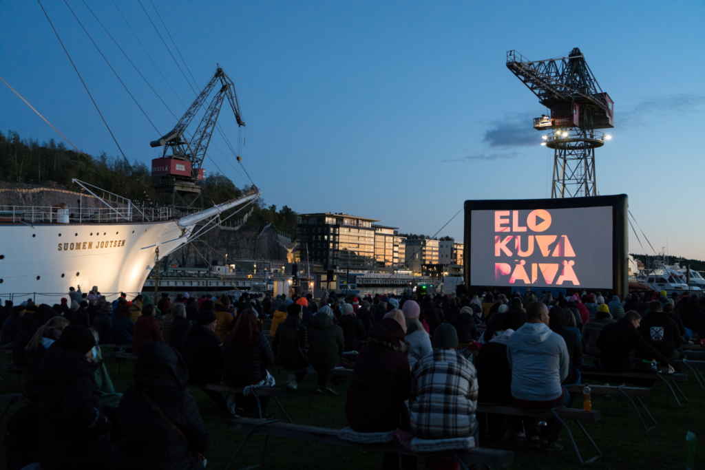 Kuva yleisön takaa, satapäinen yleisö katsoo valkokankaalta elokuvaa. Taustalla valkoinen purjelaiva, kaksi suurta telakkanosturia sekä auringonlasku.