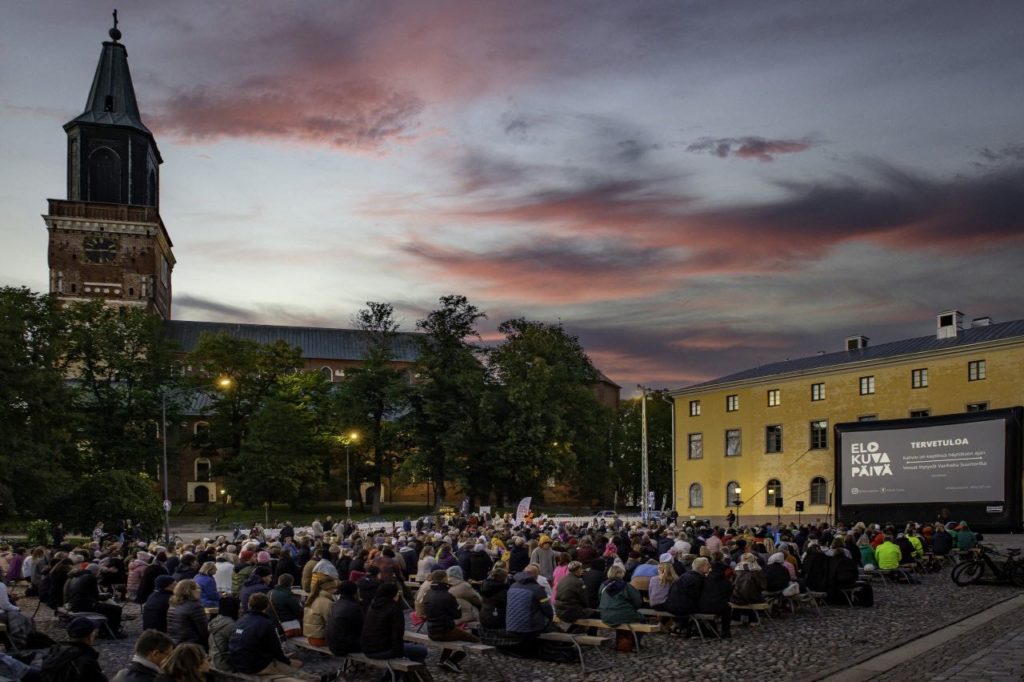 Valokuva suuresta tuhatpäisestä yleisöstä istumassa suuren kirkontornin edustalla torilla. Aurinko on laskemassa ja taivas on sinipunainen. Yleisö katsoo elokuvaa suurelta valkokankaalta, joka on pystytetty keltaisen rakennuksen eteen.