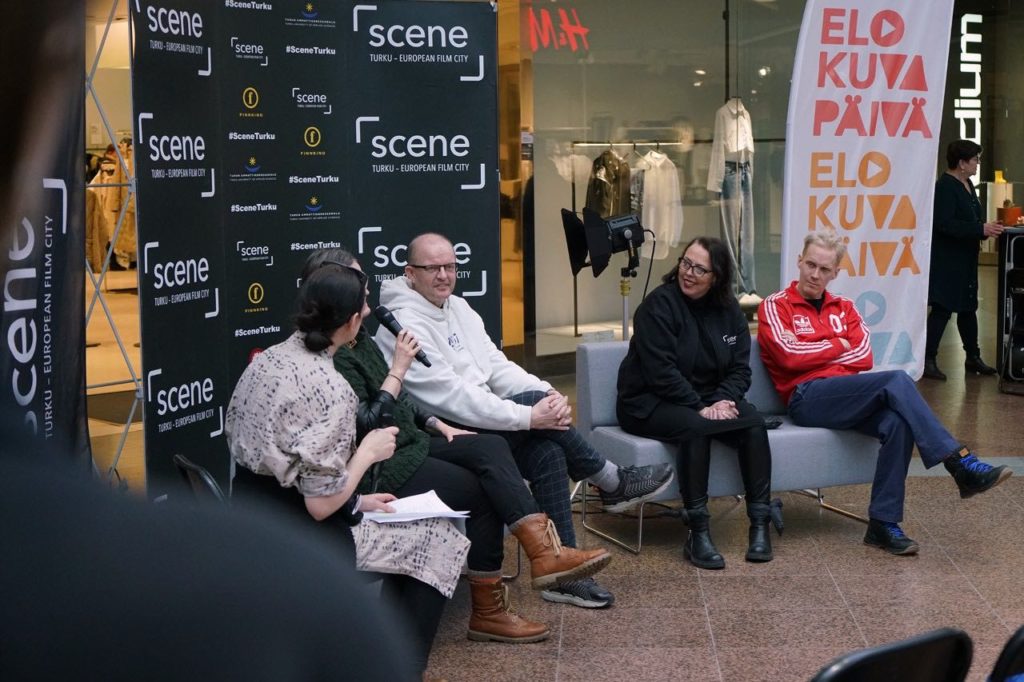 Valokuva paneelikeskustelusta, jossa neljä haastateltavaa ja yksi haastattelija istuvat harmailla sohvilla. Sohvien takana on Scenen logoilla varustettu musta seinä, sekä Elokuvapäivän liput. Paneeli on kauppakeskuksessa.